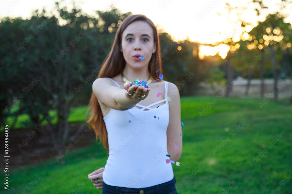 Pretty young woman standing in park on sunset and blowing confetti. Pretty young woman standing in park on sunset and blowing confetti.