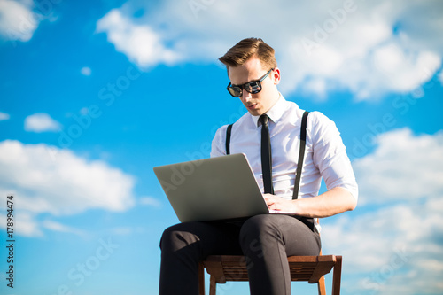 Attractive serious busunessman in white shirt, tie, braces and sunglasses work with laptop on the roof