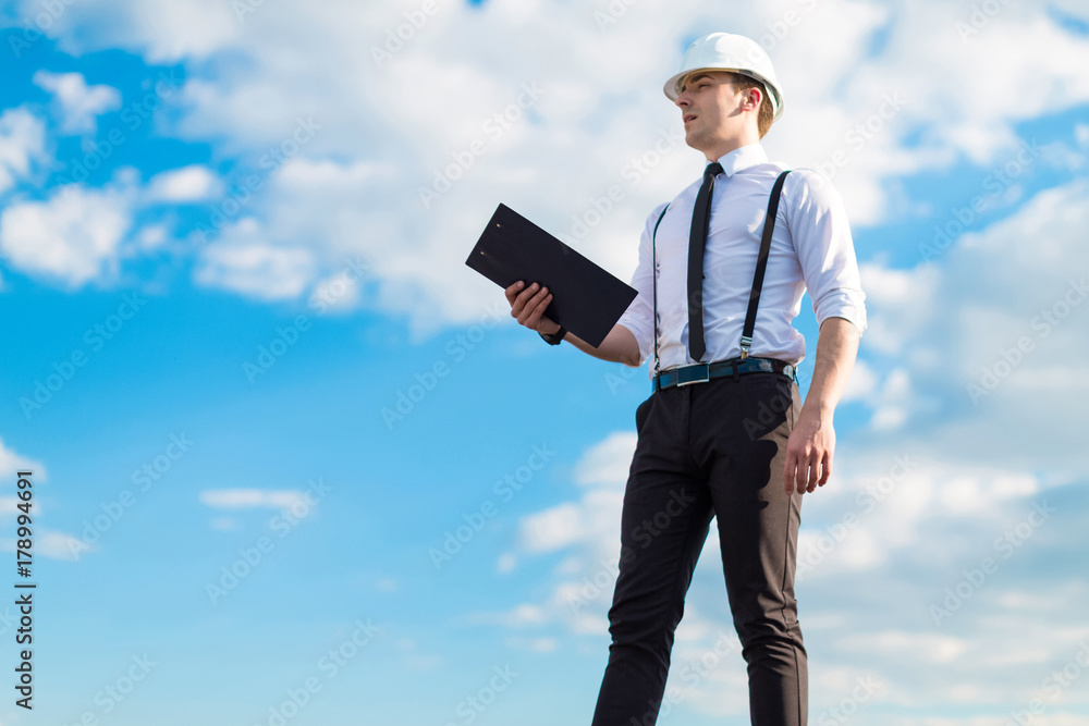 Young attractive foreman in white helmet, tie and braces stand on the roof with tablet Young attractive foreman in white helmet, tie and braces stand on the roof with tablet