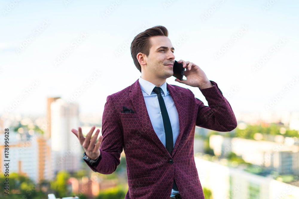 Young serious businessman in red suit and shirt with tie stand on the roof and talk on the phone Young serious businessman in red suit and shirt with tie stand on the roof and talk on the phone