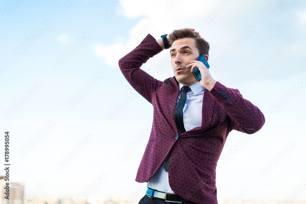 Young serious businessman in red suit and shirt with tie stand on the roof and talk on the phone Young serious businessman in red suit and shirt with tie stand on the roof and talk on the phone