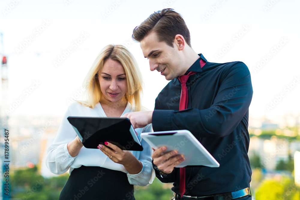 Couple of businessmen, good looking brunette man and attractive blonde woman stand on the roof and look in tablets