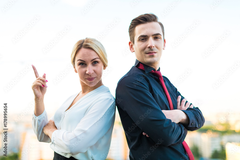 Couple of businessmen, good looking brunette man and attractive blonde woman stand on the roof back to back Couple of businessmen, good looking brunette man and attractive blonde woman stand on the roof back to back