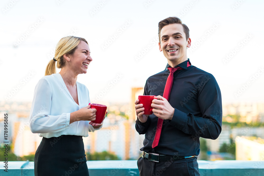 Couple of businessmen, attractive brunette man and pretty blonde woman stand on the roof and drink coffe Couple of businessmen, attractive brunette man and pretty blonde woman stand on the roof and drink coffe