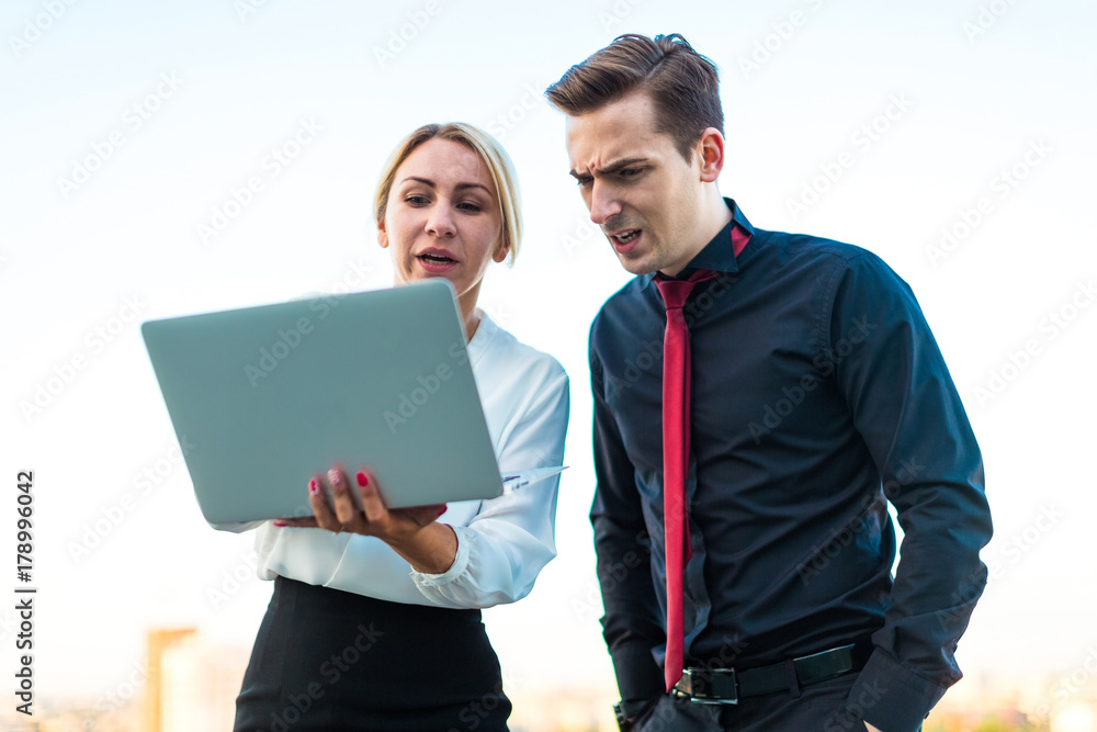 Couple of businessmen, young brunette man and young beauty blonde woman stand on the roof and look in laptop Couple of businessmen, young brunette man and young beauty blonde woman stand on the roof and look in laptop