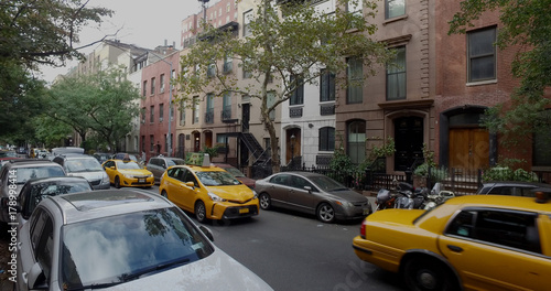 Wide view exterior shot of a typical generic New York City block with apartment buildings yellow taxi cab traffic and parked cars lining side of street.