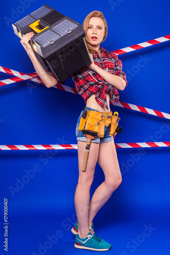 Young slim builder girl in red chechered shirt, builder belt, jean shorts and snickers hold toolbox photo