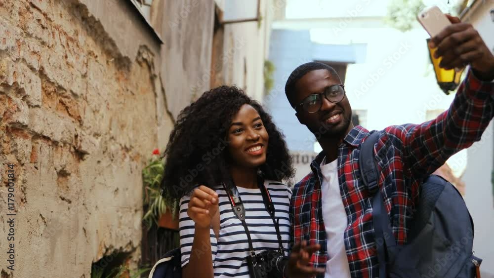 Two young tourists taking photos on mobile camera during tour in old beautiful city. Couple smiling, grimacing. Girl showing victory peace sign. Two young tourists taking photos on mobile camera during tour in old beautiful city. Couple smiling, grimacing. Girl showing victory peace sign.