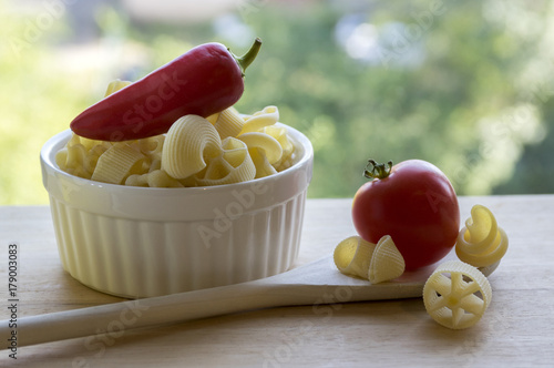 Various mix of pasta in white baking bowl on wooden table and ripened red jalapeno pepper, wooden spoon and red tomato photo