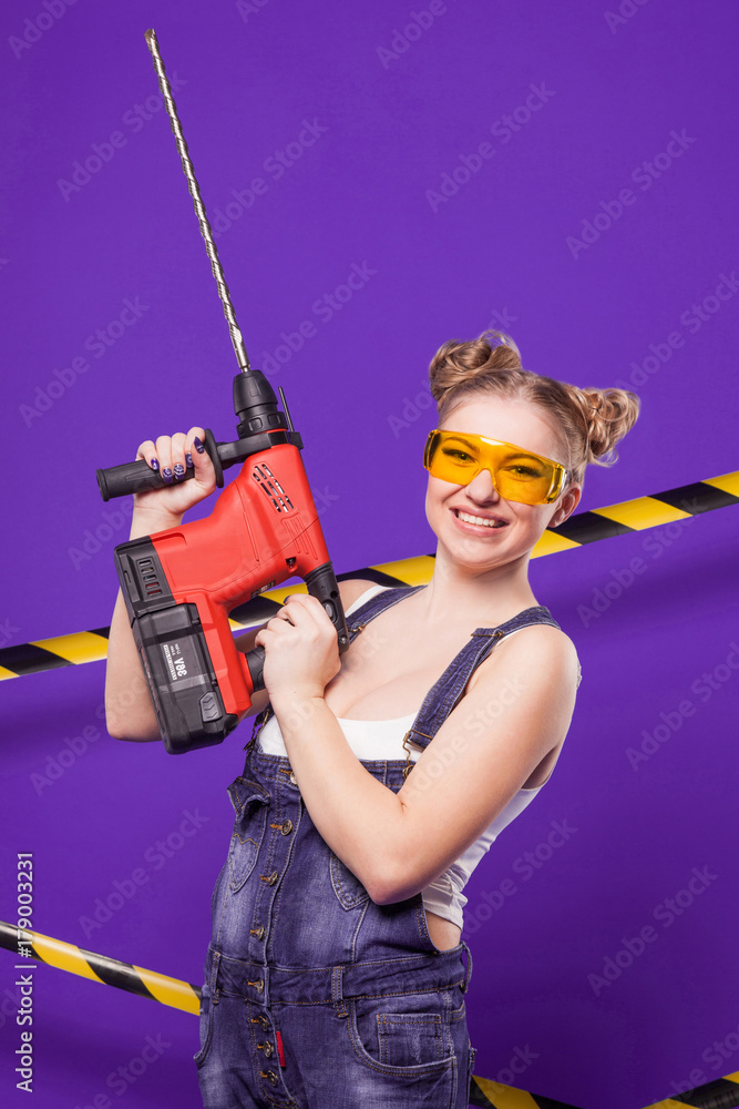 Beauty young builder girl in denim jumpsuit and glasses stand with big drill