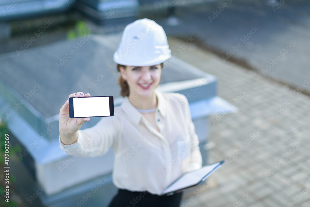 Attractive businesslady in white blouse, watch, helmet and black skirt stand on the roof and show empty smartphone