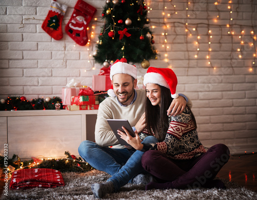 Adorable young couple sitting on a carpet with Santa hats hugged while looking on a tablet for Chrismas holidays.