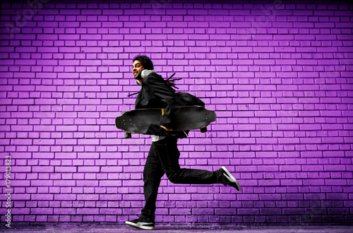 Stylish happy dreadlocks skater with headphones running in a suit near the violet wall on the street.