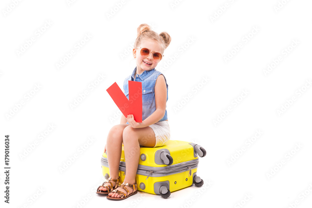 Attractive little girl in blue shirt, white shorts and sunglasses sit on the yellow suitcase and hold red cards Attractive little girl in blue shirt, white shorts and sunglasses sit on the yellow suitcase and hold red cards