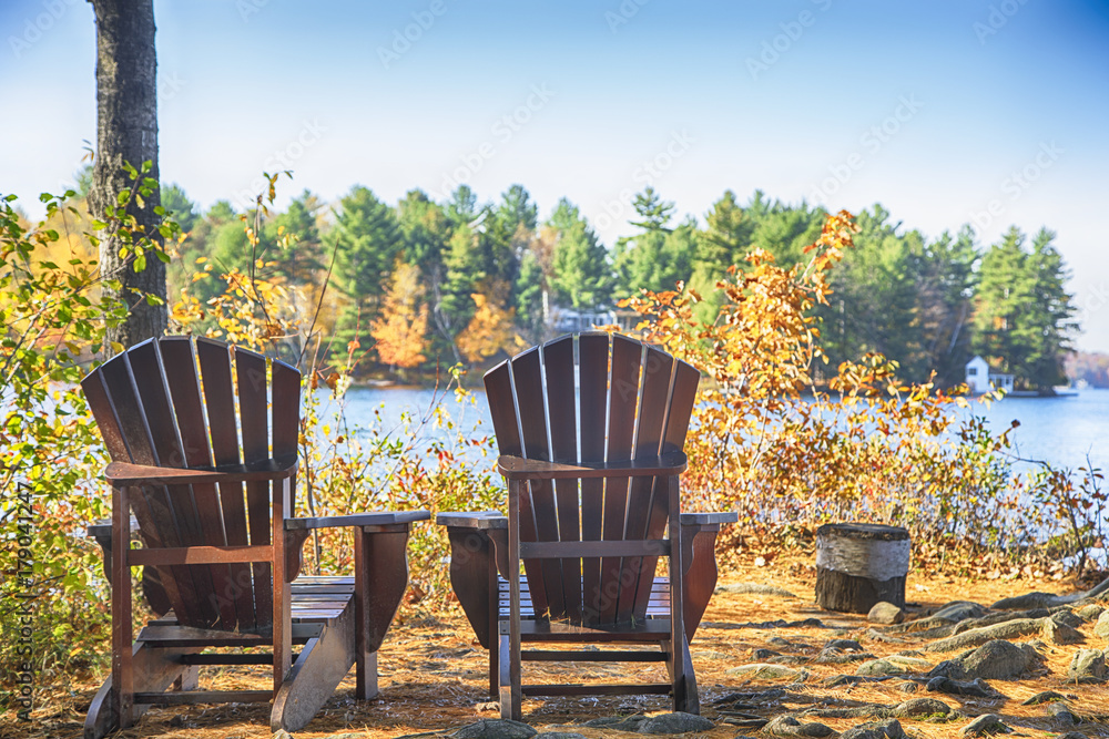 Naklejka premium Two Muskoka chairs on a wood dock at a blue lake