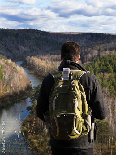 Outdoor portrait from back. A man in a black coat and a backpack looks at the river among the mountains.