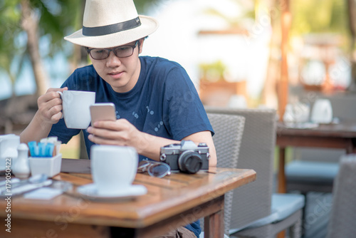 Young Asian man with a cup of coffee using smart phone during having breakfast in resort, breakfast time and summer holiday vacation concepts