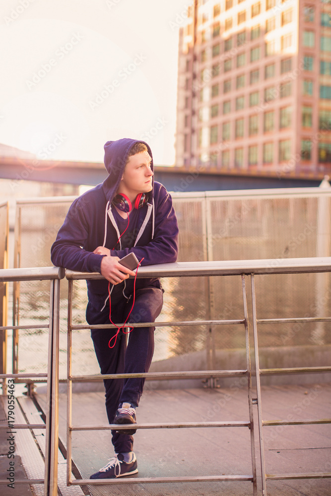 The young sporty man standing outdoor in the earphones and holding the phone in hand and looking aside