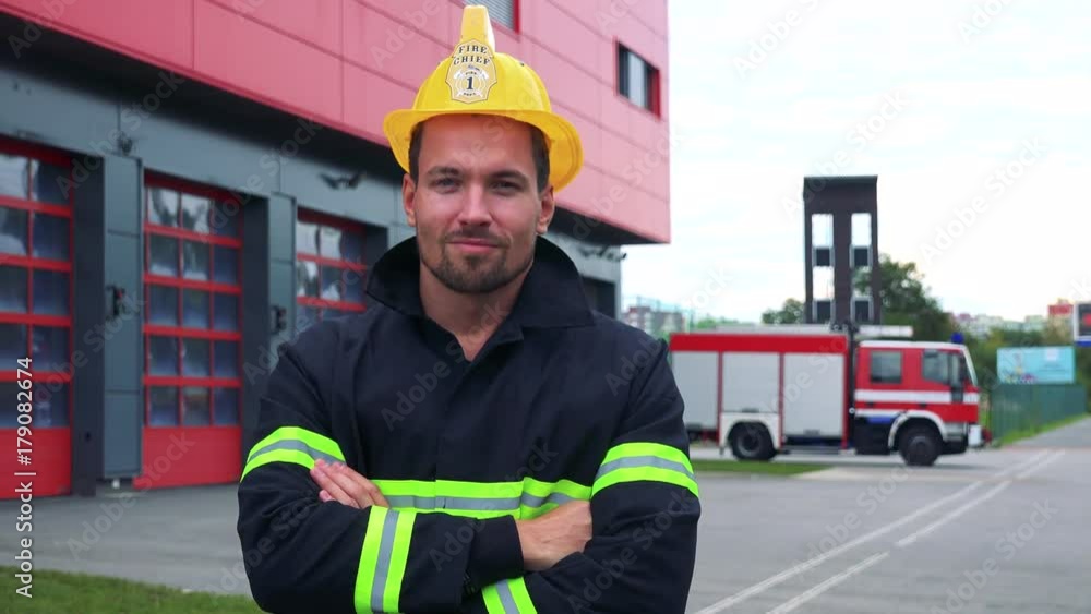 A young firefighter looks at the camera with arms folded across chest - a truck drives into a fire station garage in the background A young firefighter looks at the camera with arms folded across chest - a truck drives into a fire station garage in the background
