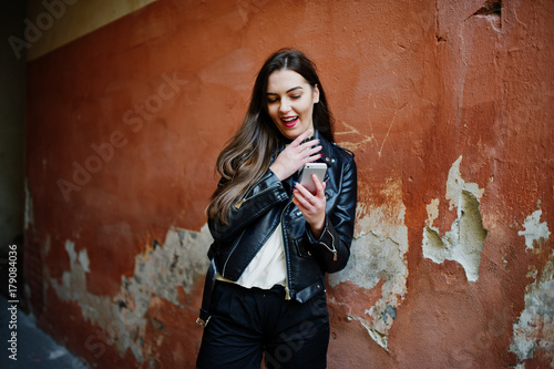 Stylish brunette girl wear on leather jacket and shorts with mobile phone at hands against orange wall. © AS Photo Family