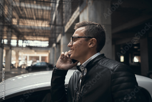 Close-up. A young handsome man talking on the phone. He smiles. Standing at the car parking