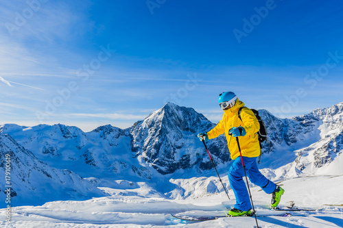 Mountaineer backcountry ski walking up along a snowy ridge with skis in the backpack. In background blue sky and shiny sun and Zebru, Ortler in South Tirol, Italy. Adventure winter extreme sport.