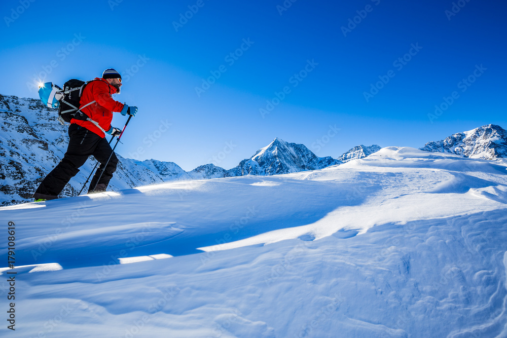 Mountaineer backcountry ski walking up along a snowy ridge with skis in the backpack. In background blue sky and shiny sun and Zebru, Ortler in South Tirol, Italy.  Adventure winter extreme sport.