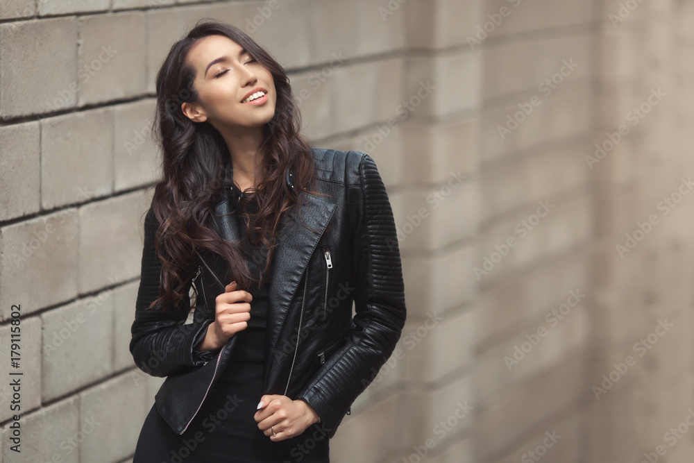 Portrait of beautiful smiling brunette, fashion and stylish standing near the wall. A curly hairstyle woman in total black look with leather jacket and dress. Portrait of beautiful smiling brunette, fashion and stylish standing near the wall. A curly hairstyle woman in total black look with leather jacket and dress.