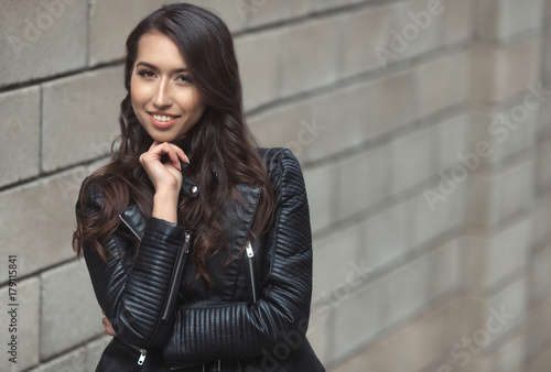 Portrait of beautiful smiling brunette, fashion and stylish standing near the wall. A curly hairstyle woman in total black look with leather jacket and dress.