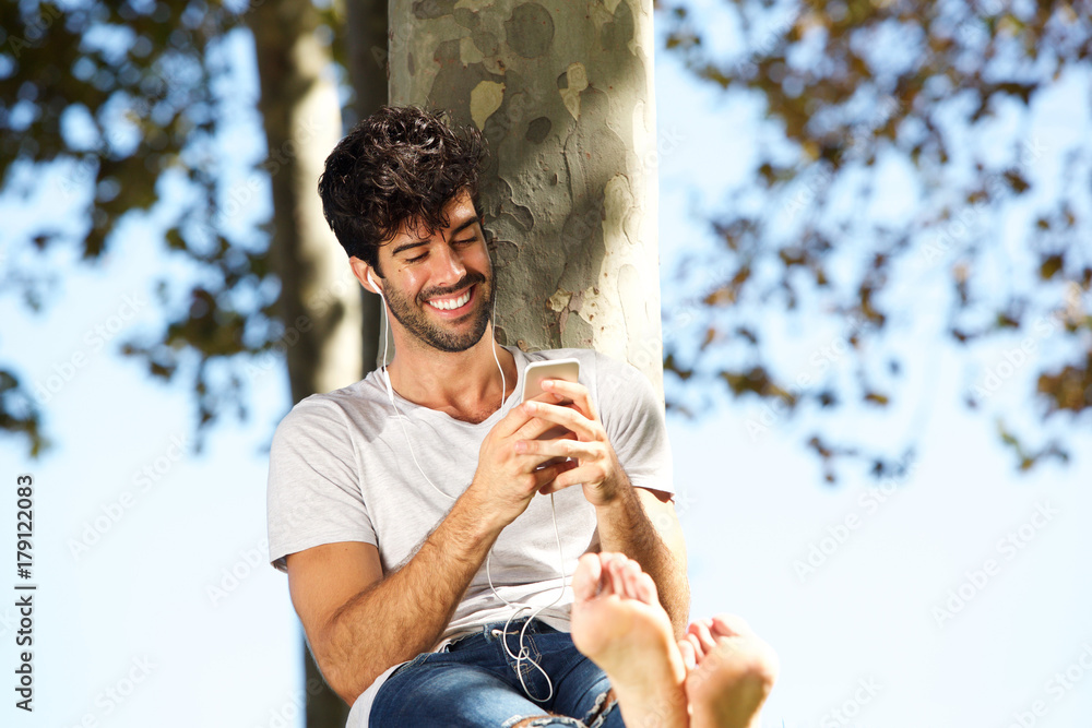 Fototapeta premium Smiling man sitting by tree barefoot with headphones and cellphone