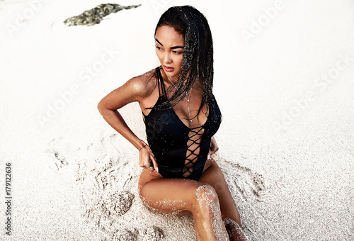 portrait of beautiful caucasian sunbathed woman model with dark long hair in black swimsuit posing on summer beach with white sand