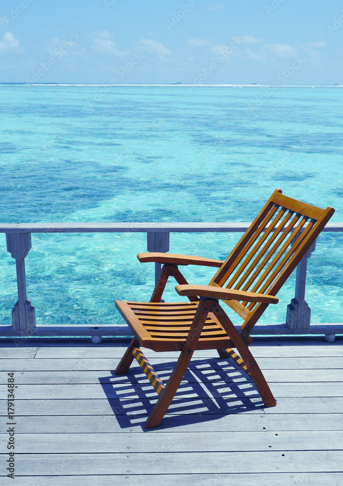 Chair near ocean Tropical Holiday Banner. One deck chair on a sea view balcony at Maldives. One chaise longue at the beach with clear skies and blue ocean. Maldives luxury vacation on the holidays.