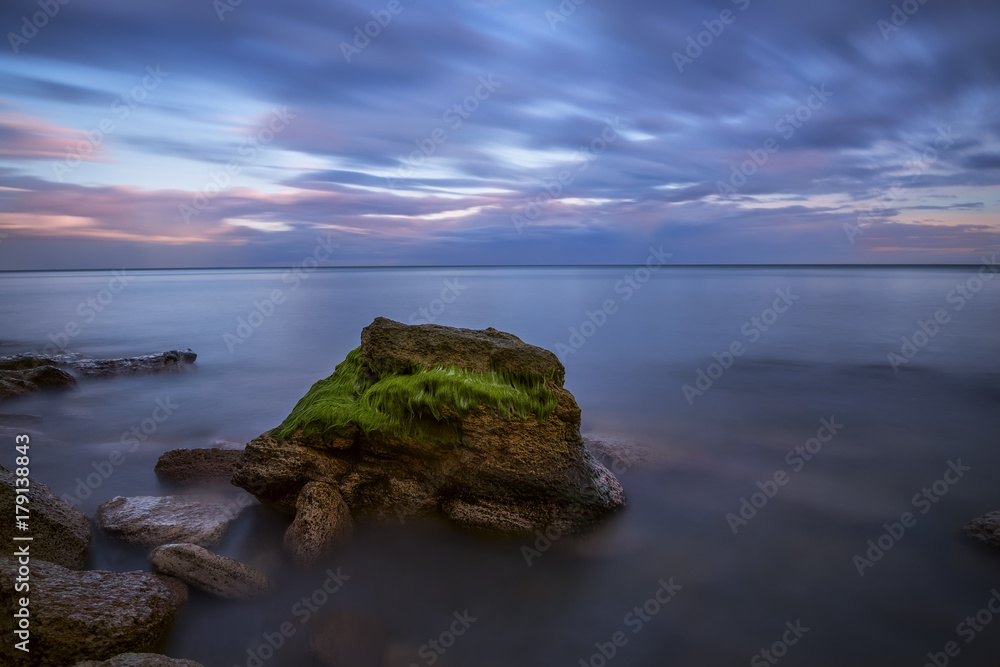 Sea landscape at sunset. The blurred surface of the water on a long exposure, a stone covered with green moss and a beautiful sky with running blurry clouds. Calm evening marine look.
