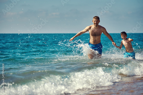 Caucasian dad and son are running in the water along the sea shore. They are holding hands. © Artem