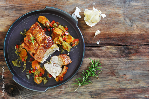Crispy roast pork with vegetables, rosemary and garlic on a rustic dark wooden table, top view from above, copy space