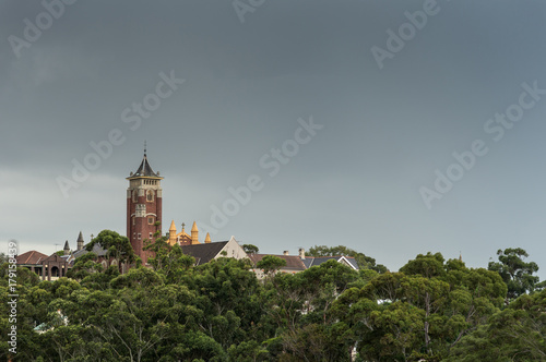 Sydney, Australia - March 21, 2017: The brown, square spire, crosses and roofs of Saint Augustine Catholic Church towers over the green park in front under a rainy gray sky.