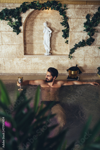 Young handsome bearded man enjoying and relaxing in spa center. He sitting in Jacuzzi or hot tub and drinking wine. © Dusko