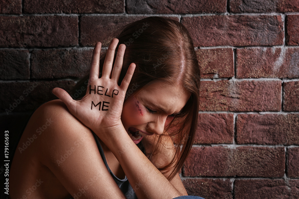 Φωτογραφία Battered young woman with written words Help me on palm near brick wall