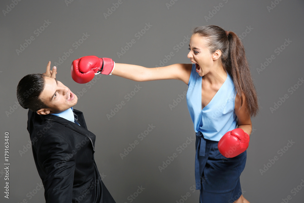 Woman in formal clothes and boxing gloves hitting businessman on grey background Woman in formal clothes and boxing gloves hitting businessman on grey background