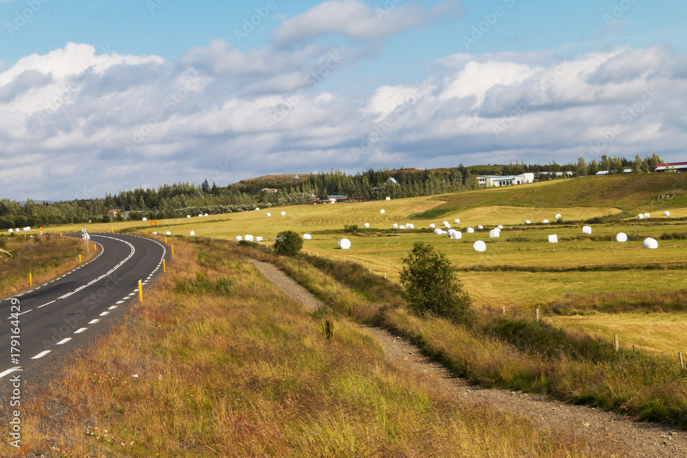 Iceland - typical landscape with roadway, mountains, baled silage, hay, green grass. blue sky, farm houses and clouds.