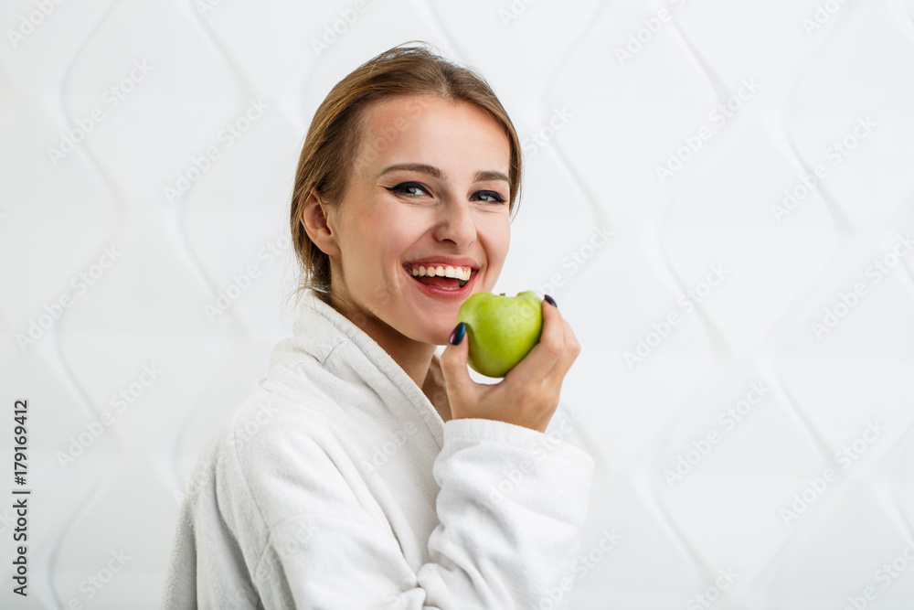 Cheerful woman in the white bathrobe enjoys a green apple, indoor shot in the white background Cheerful woman in the white bathrobe enjoys a green apple, indoor shot in the white background