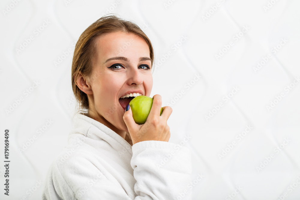 Cheerful woman in the white bathrobe enjoys a green apple, indoor shot in the white background Cheerful woman in the white bathrobe enjoys a green apple, indoor shot in the white background