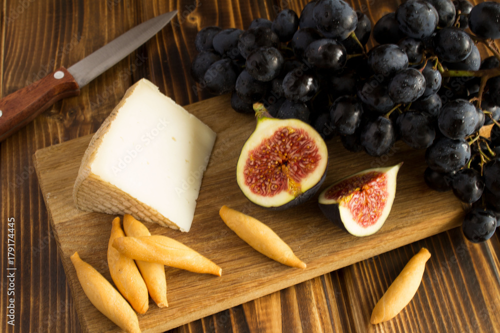 Piece of cheese, fruits and bread sticks on the brown cutting board