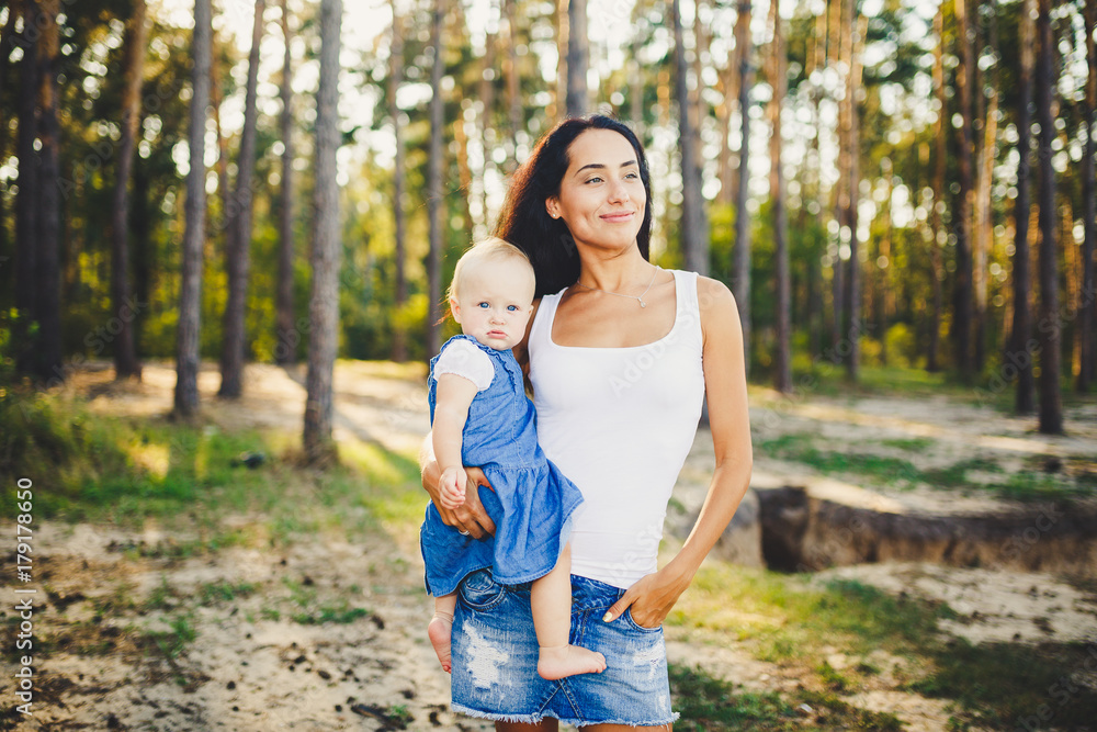 Fototapeta premium young beautiful fashionable mother with long brunette hair holding a daughter's blonde with blue eyes one year of birth in a coniferous forest in a summer park.