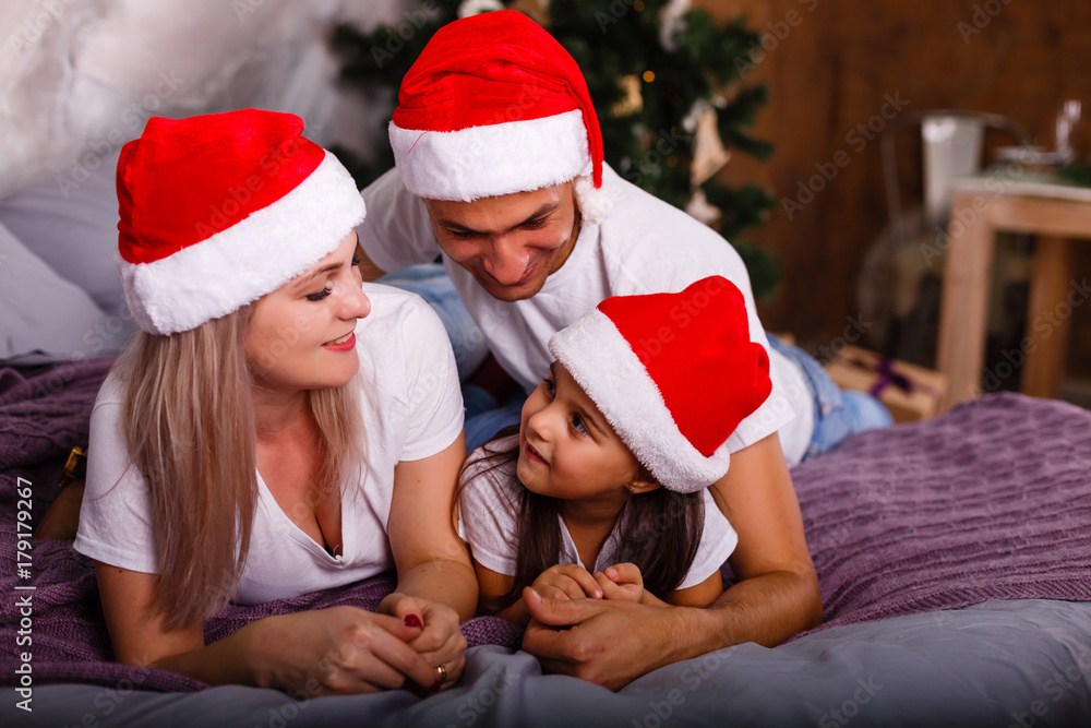 Fototapeta premium Family in Christmas Santa hats lying on bed. Mother; father and baby having fun in bedroom. People relaxing at home. Winter holiday Xmas and New Year concept
