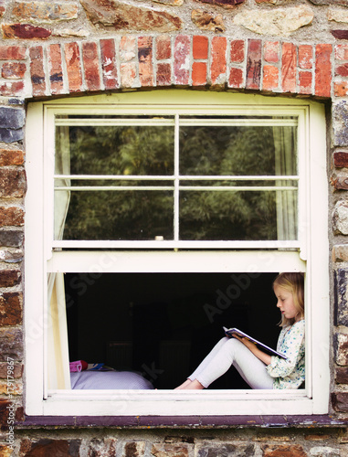 A little girl sitting a window reading a book. photo