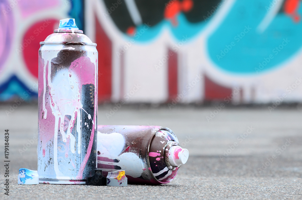Naklejka premium Kilka zużytych puszek natryskowych z różową i białą farbą oraz nakrętek do rozpylania farby pod ciśnieniem leży na asfalcie w pobliżu pomalowanej ściany w kolorowych rysunkach graffiti