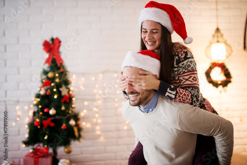 Close up of attractive playful love couple with Santa hats and sweaters having a piggyback ride for Christmas holidays in the living room.
