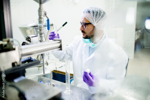 Young bearded scientist with gloves and hair protection checking test tube at laboratory table.