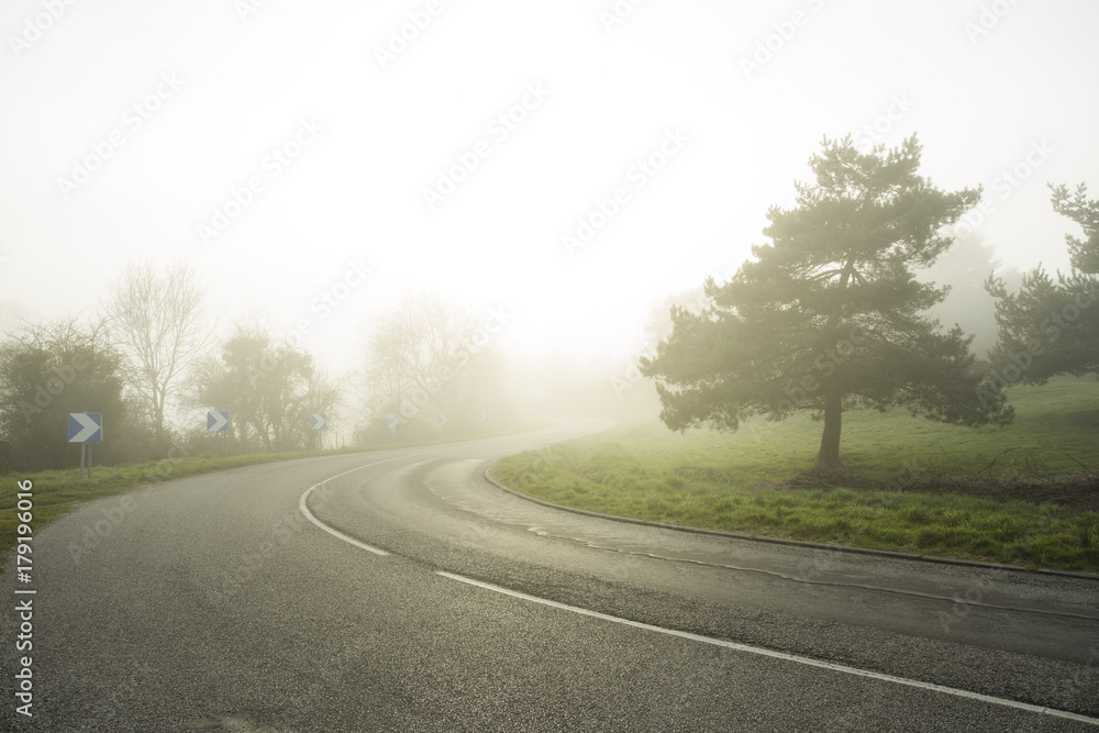 Foggy asphalt curved road passing through the forest. Weather with low visibility in the region of Normandy, France. Country landscape on misty day. Toned Foggy asphalt curved road passing through the forest. Weather with low visibility in the region of Normandy, France. Country landscape on misty day. Toned